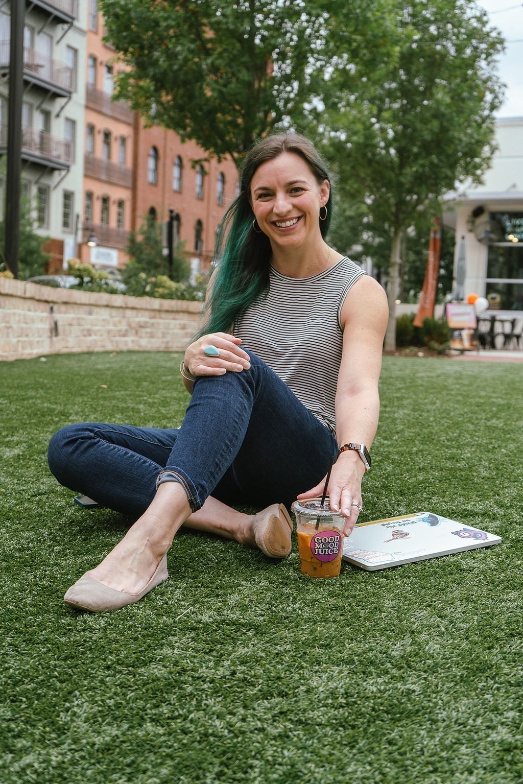 Young woman sitting in grass in an open suburban area with laptop and coffee