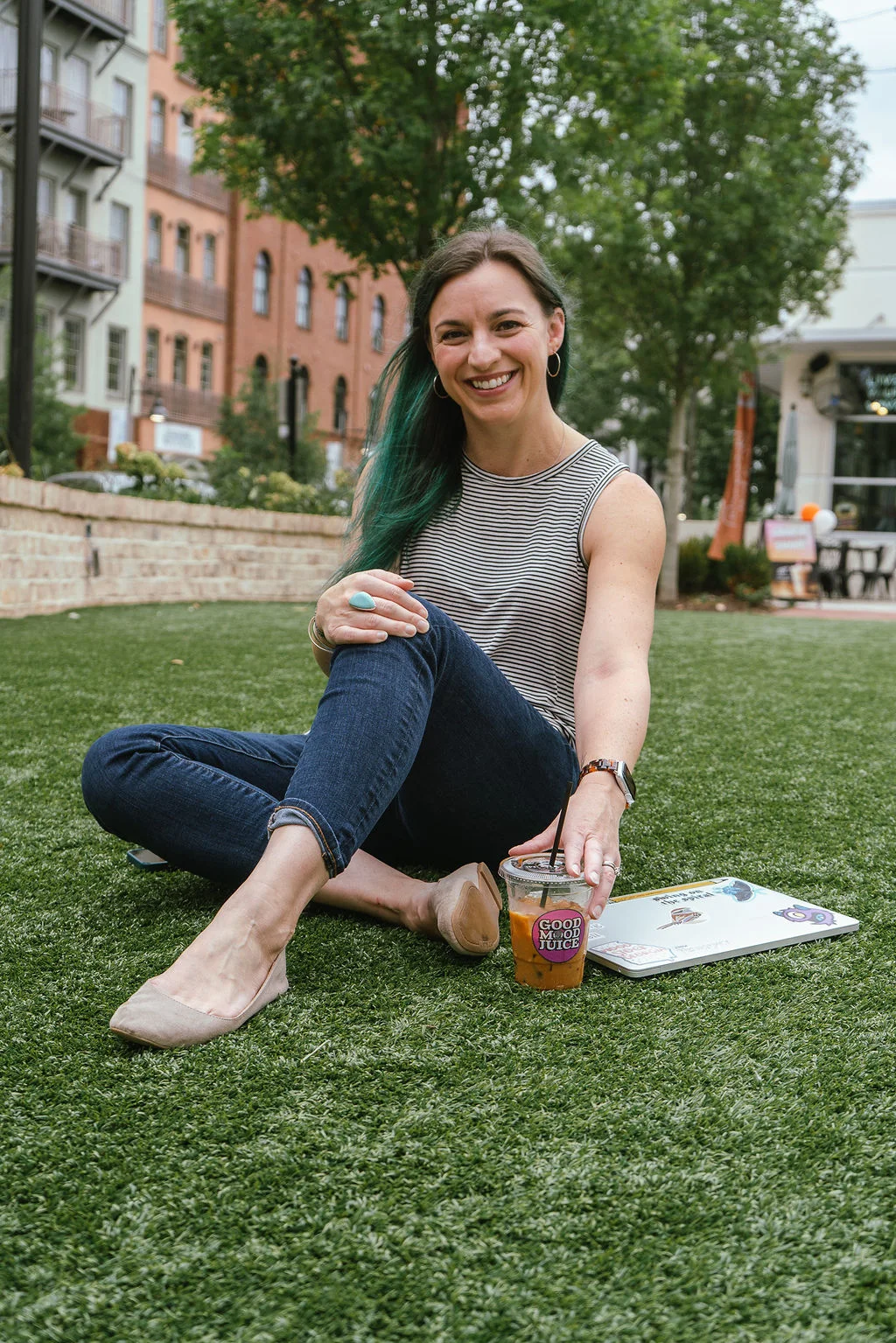 Young woman sitting in grass in an open suburban area with laptop and coffee